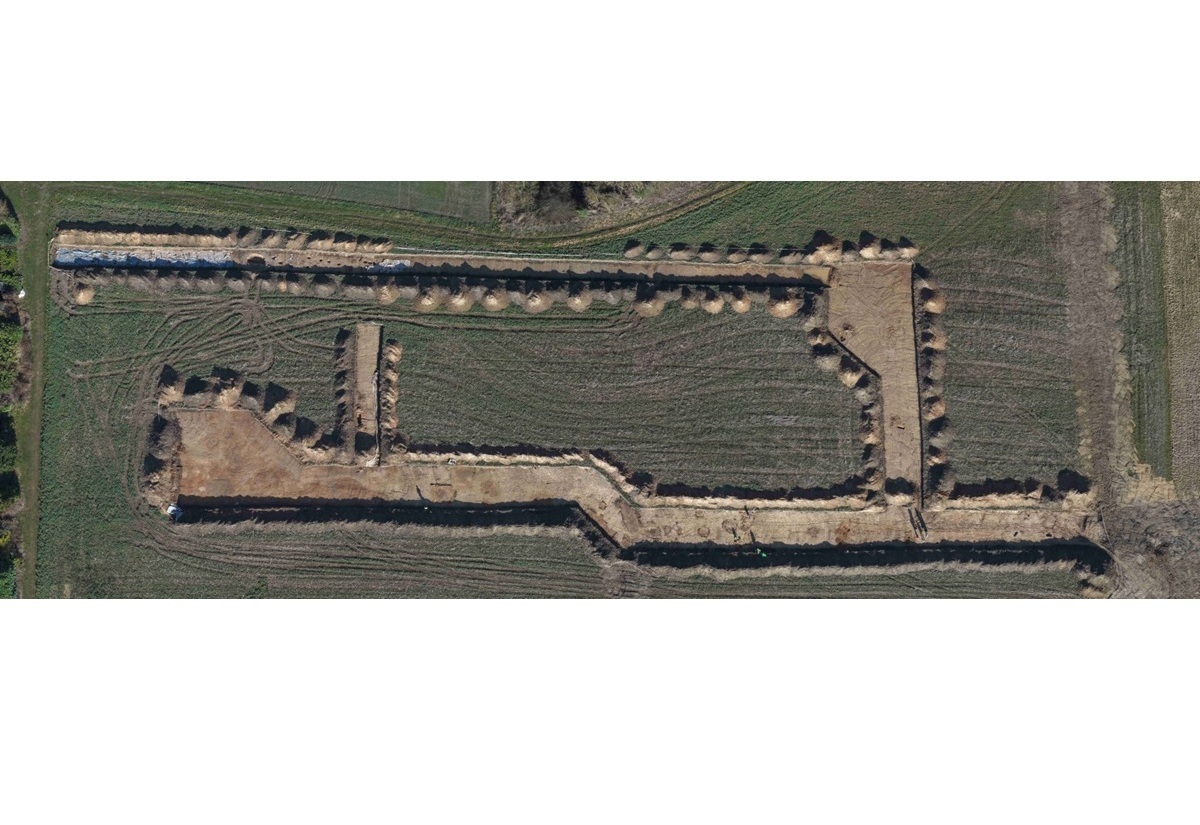 Luftaufnahme eines rechteckigen Ausgrabungsfelds mit klar abgegrenzten, hellen Erdflächen, die von dunklerem, bewachsenem Boden umgeben sind. Die freigelegten Bereiche verlaufen in geraden und abgewinkelten Linien und zeigen mehrere parallele und rechtwinklige Strukturen. Die Umgebung besteht aus grün-braunen Feldern mit sichtbaren Traktorspuren.