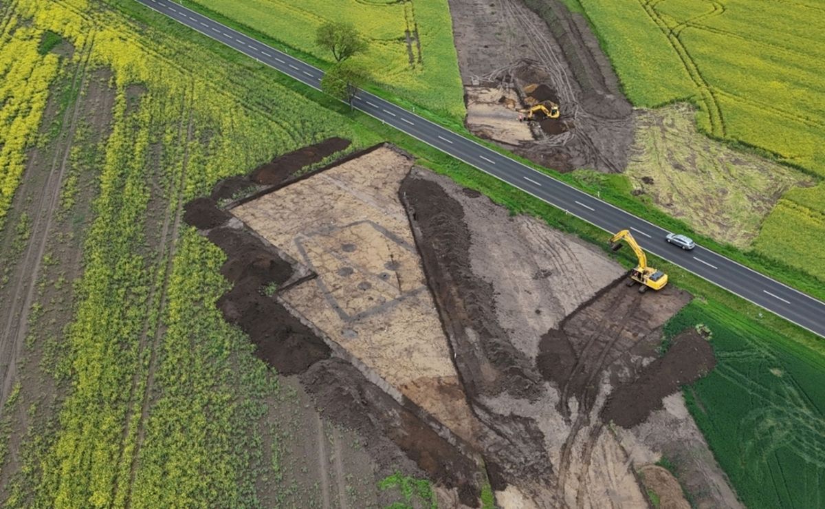 Luftaufnahme einer trapezförmigen archäologischen Ausgrabungsstätte neben einer Landstraße mit zwei Baggern und umliegenden Feldern