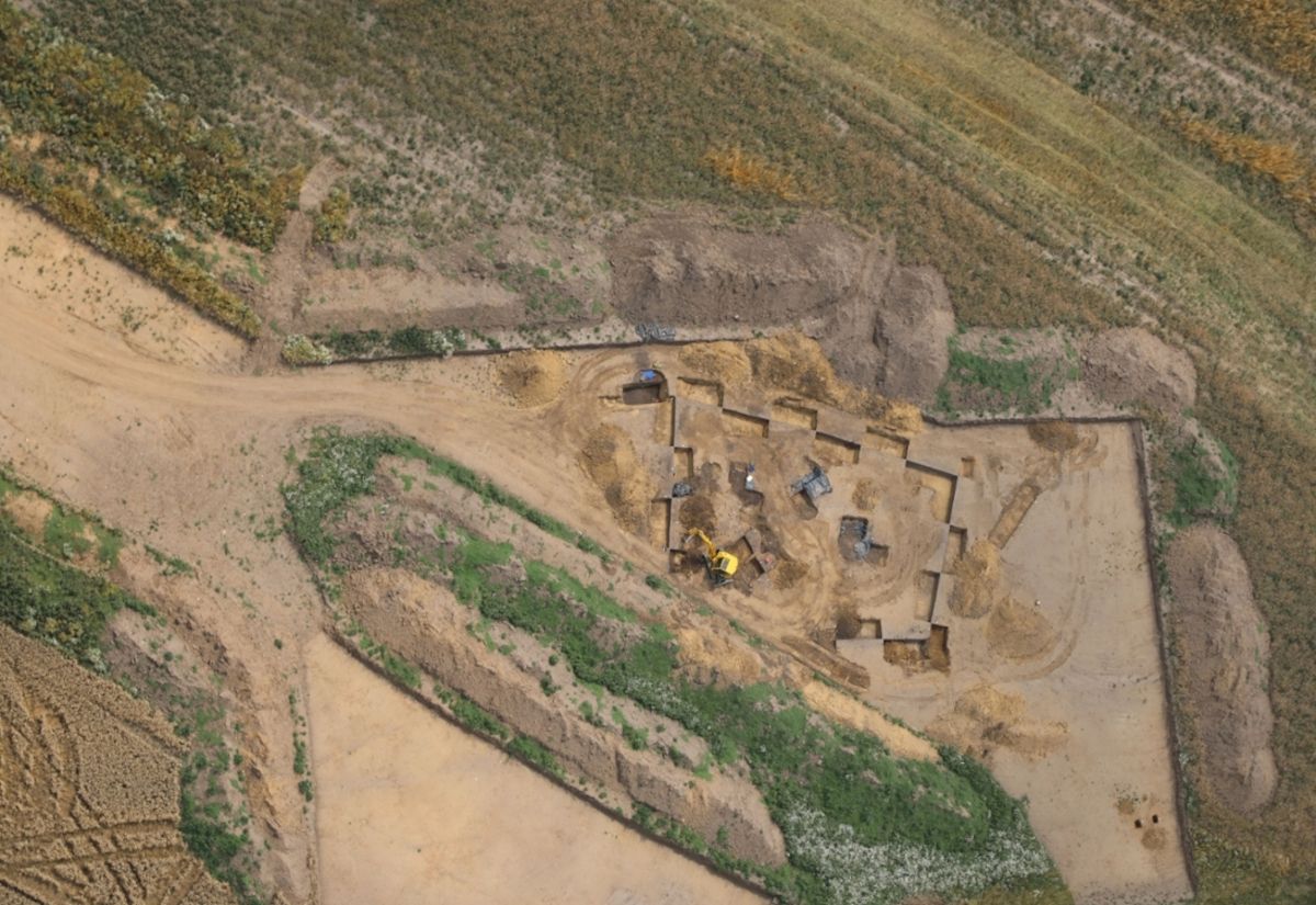 Luftaufnahme einer trapezförmigen Ausgrabungsstätte aus der Jungsteinzeit mit mehreren rechteckigen Gruben und einem Bagger in einer ländlichen Umgebung. Landkreis Leipzig
