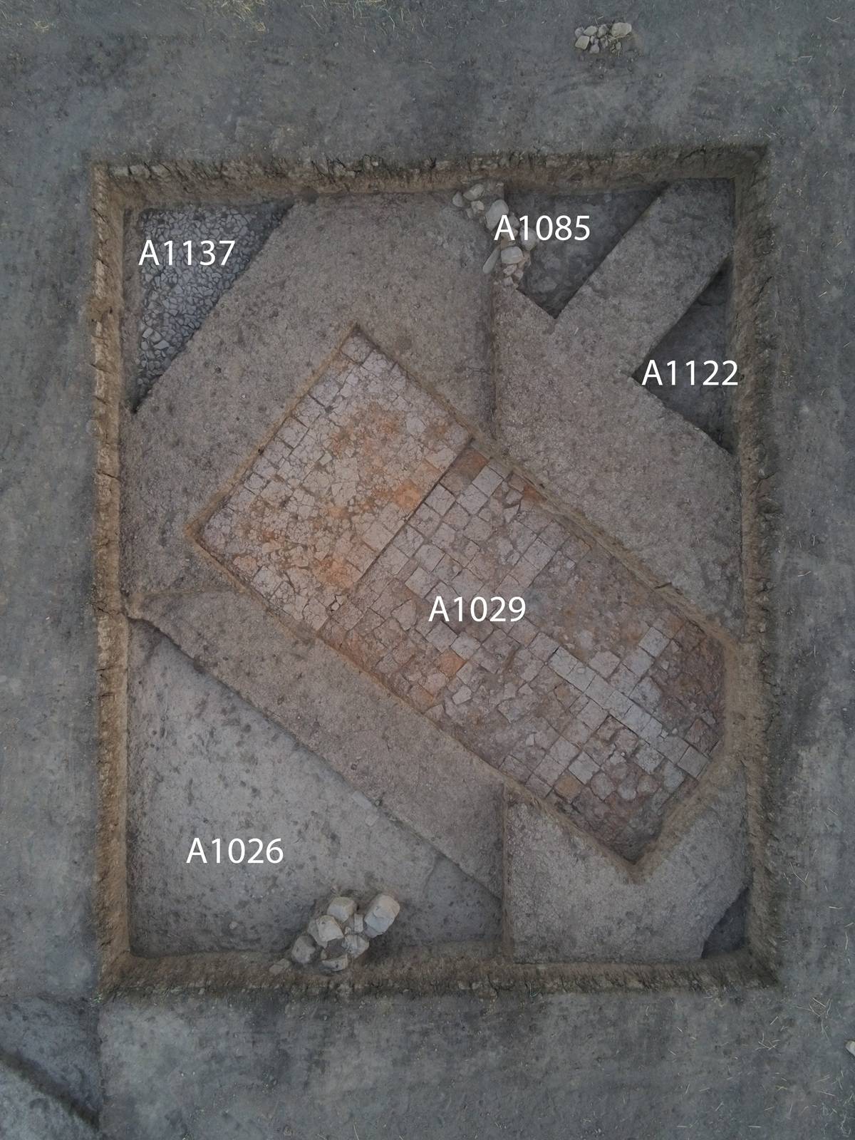 Archäologische Ausgrabungsstätte mit rechteckigem, von Erde umgebenem Bereich, der in mehrere Abschnitte unterteilt ist. Innerhalb des Bereichs sind unterschiedliche Bodenbeläge sichtbar, darunter ein zentraler rechteckiger Abschnitt mit quadratischen Ziegeln. Die Abschnitte sind mit den Bezeichnungen 'A1026', 'A1029', 'A1085', 'A1122' und 'A1137' markiert.