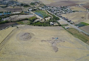 Luftaufnahme eines archäologischen Ausgrabungsgeländes in einer ländlichen Gegend. Im Vordergrund sind zwei größere Ausgrabungsstellen mit sichtbaren Erdschichten und Grabungsgruben zu sehen. Im Hintergrund erstreckt sich ein Dorf mit mehreren Gebäuden, Straßen und landwirtschaftlichen Feldern. Die Landschaft ist überwiegend flach und von Feldern und vereinzelten Bäumen geprägt.