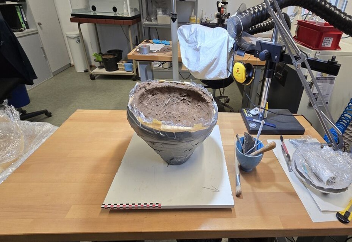 Im Mittelpunkt steht ein großer, halbkugelförmiger Erdblock, der mit Folie und Klebeband gesichert ist. Er befindet sich auf einer weißen Platte auf einem Holztisch. Daneben liegen archäologische Werkzeuge wie Pinsel, Spatel und ein kleiner Mörser. Im Hintergrund sind Labormöbel, Geräte, Schubladen und weitere Arbeitsmaterialien zu sehen. Die Szene spielt in einem modernen Laborraum.