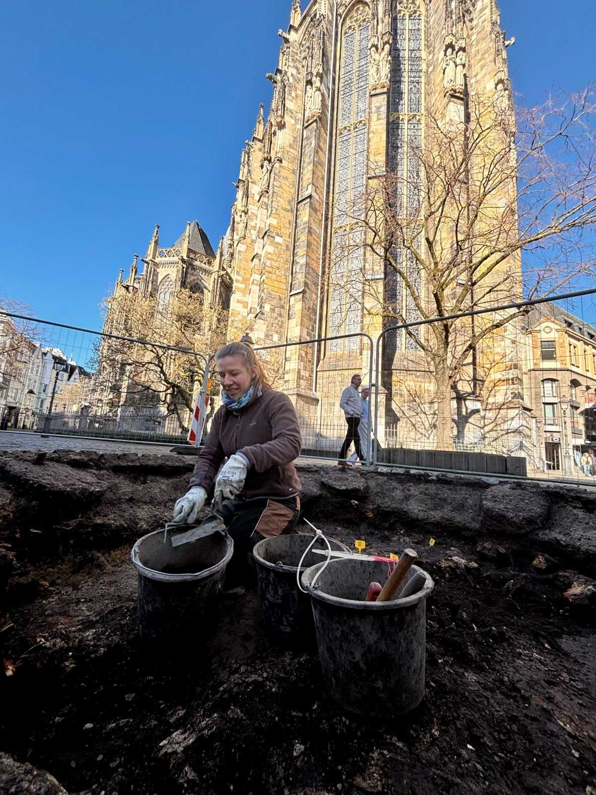 Im Vordergrund kniet eine Person in dunkler Arbeitskleidung und Handschuhen in einer Ausgrabungsgrube. Vor ihr stehen mehrere Eimer und Grabungswerkzeuge auf dunklem Erdreich. Im Hintergrund erhebt sich die monumentale Fassade eines gotischen Gebäudes mit hohen Fenstern und detaillierten Steinstrukturen. Ein Baum ohne Laub und weitere Gebäude sind sichtbar. Der Himmel ist klar und blau.