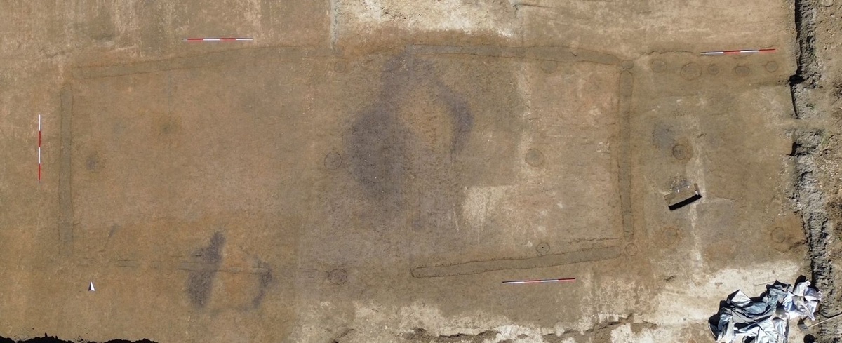 Rechteckige Ausgrabungsfläche mit deutlich sichtbaren, dunkleren Verfärbungen im Boden, die eine größere rechteckige Struktur und mehrere kleinere, rundliche Bereiche markieren. Zwei rot-weiße Messlatten liegen parallel an gegenüberliegenden Seiten der Fläche. Am rechten Rand sind helle, gestapelte Säcke zu erkennen.