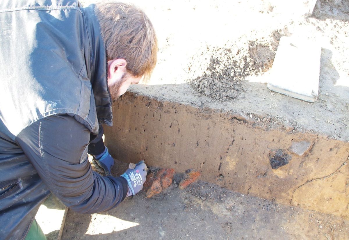 Eine Person mit Handschuhen kniet an einer Ausgrabungsstelle und arbeitet vorsichtig an einer Erdschicht. Im Profil sind orangefarbene Fragmente im Boden sichtbar, die sich deutlich vom umgebenden Erdreich abheben. Die Szene ist von Tageslicht erhellt, der Boden wirkt trocken und sandig. Im Hintergrund liegen weitere Erdklumpen und ein heller rechteckiger Gegenstand.