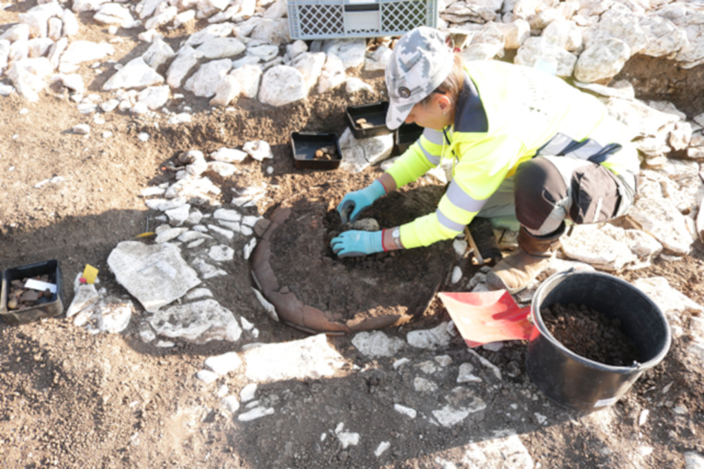 Eine Person mit gelber Warnjacke, heller Hose und gemustertem Hut kniet auf dem Boden und arbeitet mit blauen Handschuhen an einer Ausgrabungsstelle. Im Vordergrund ist ein halbkreisförmiger, steinerner Rand im Erdreich freigelegt. Um die Person liegen Werkzeuge, ein schwarzer Eimer und eine Kiste. Der Boden ist teilweise mit Steinen bedeckt.
