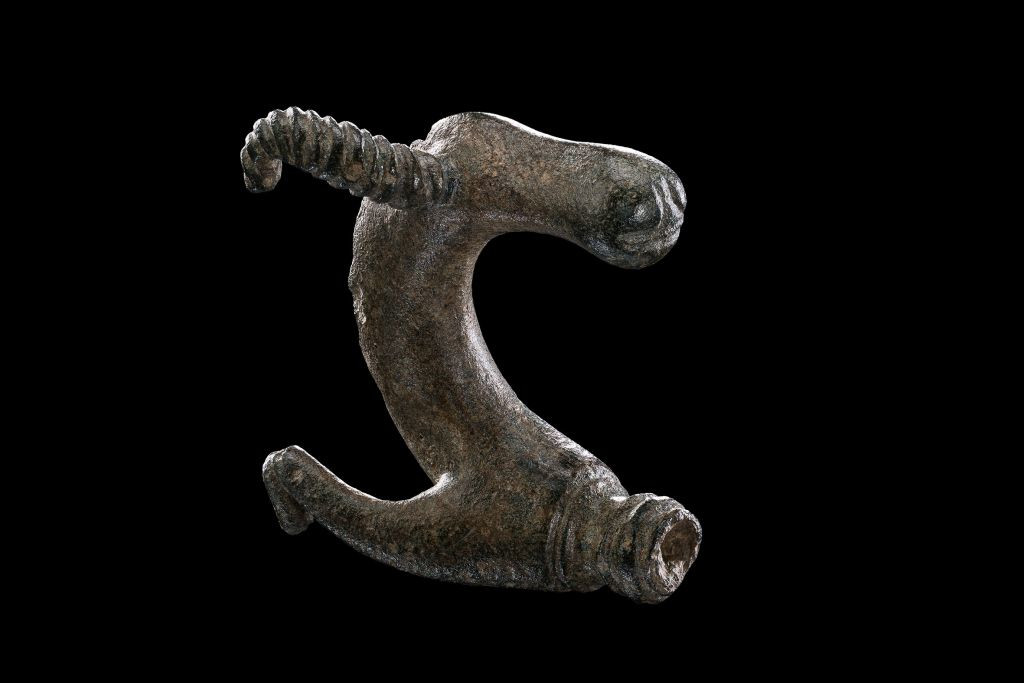 Gebogener Metallgegenstand mit rauer Oberfläche und zwei unterschiedlich gestalteten Enden. Das eine Ende ist spiralförmig gewunden, das andere leicht geöffnet und glatt. Die Form erinnert an einen Haken oder eine Spange. Der Gegenstand liegt vor schwarzem Hintergrund.