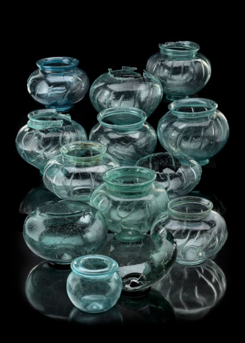 Mehrere bauchige Glasgefäße mit breitem Hals und transparentem, leicht grünlichem Glas stehen auf einer schwarzen Fläche. Einige Gefäße sind beschädigt oder weisen Risse auf. Die Gefäße sind unterschiedlich groß und wirken antik
