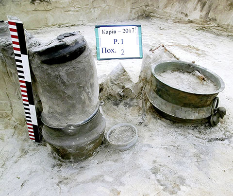 Zwei große Metallgefäße stehen nebeneinander auf sandigem Boden. Das linke Gefäß ist zylindrisch und wirkt massiv, das rechte ist flacher und rund. Beide zeigen deutliche Gebrauchsspuren und sind teilweise von Erde bedeckt. Links neben den Gefäßen steht eine vertikale Messlatte mit abwechselnd schwarzen, weißen und roten Streifen. Im Hintergrund befindet sich ein rechteckiges Schild mit kyrillischer Schrift und Zahlen.