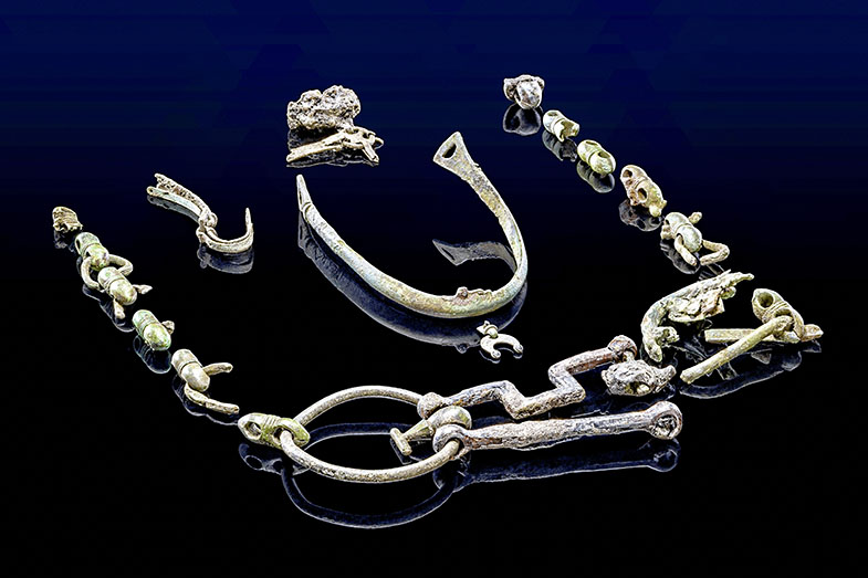 Mehrere Metallobjekte liegen auf einer glänzenden, dunkelblauen Oberfläche. Die Gegenstände sind unterschiedlich geformt und weisen Korrosionsspuren sowie Ablagerungen auf. Einige Teile sind miteinander verbunden, andere liegen lose. Die Objekte zeigen verschiedene Strukturen, darunter Ringe, Haken und kleine, kugelförmige Elemente.