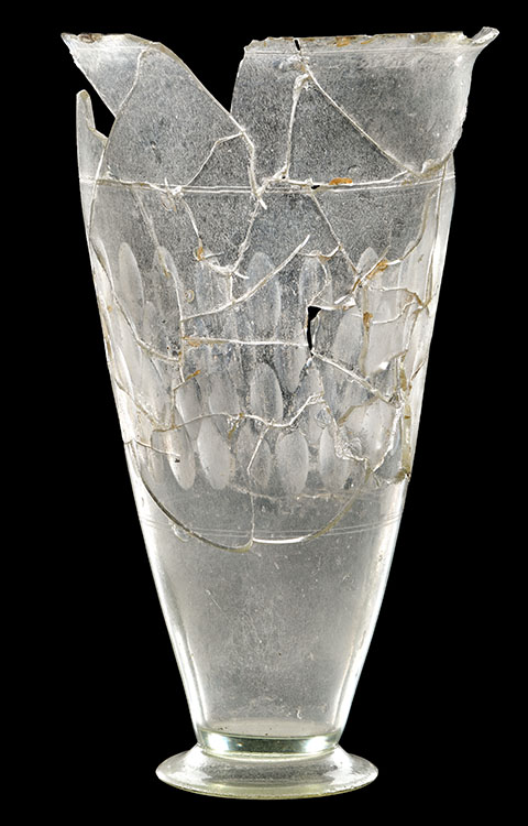 Ein hohes, schlankes Glasgefäß mit breitem Fuß und schmaler Öffnung. Die Oberfläche zeigt zahlreiche Risse und Bruchstellen, die mit feinen Linien sichtbar sind. Das Gefäß ist transparent und weist eine ovale Musterung auf, die sich in mehreren Reihen an der Außenseite abzeichnet. Der obere Rand ist unregelmäßig und teilweise abgebrochen.