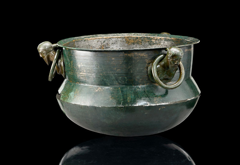 Ein bronzener Kessel mit grünlicher Patina steht auf schwarzem Hintergrund. An zwei gegenüberliegenden Seiten sind plastische Köpfe angebracht, um ihren Hals jeweils ein großer Ring. Die Oberfläche zeigt Gebrauchsspuren und leichte Korrosion.