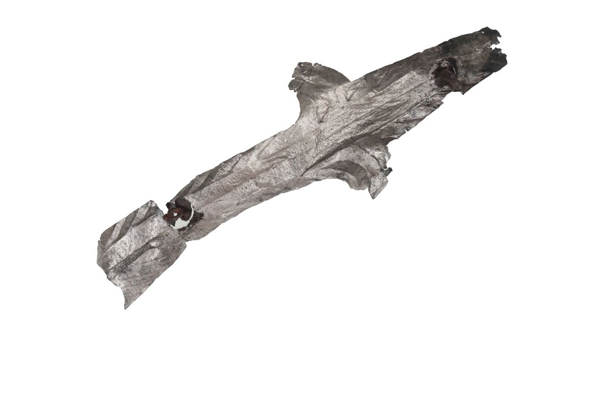 Ein länglicher, unregelmäßig geformter Gegenstand mit mehreren Ausbuchtungen und dunklen Stellen liegt auf weißem Hintergrund. Die Oberfläche wirkt rau und strukturiert, an mehreren Stellen sind Vertiefungen oder Löcher zu erkennen. Die Farbgebung ist überwiegend grau mit dunkleren Akzenten.