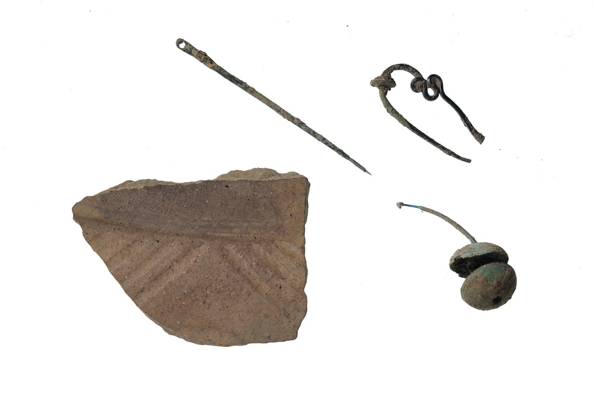 Vier archäologische Fundstücke: ein spitz zulaufender Metallstift, ein gebogener Draht mit Schlaufen, ein rundes Metallobjekt mit Stiel und ein Scherbenfragment mit Rillenmuster