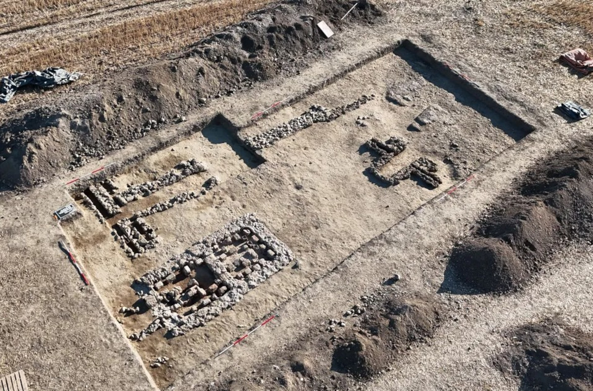 Luftaufnahme einer archäologischen Ausgrabungsstätte mit rechteckigem Grundriss. Im Zentrum und am Rand verlaufen freigelegte Mauerreste aus Stein, die verschiedene Räume und Strukturen andeuten. Der Boden ist trocken und hell, umliegende Erdschichten sind abgetragen.