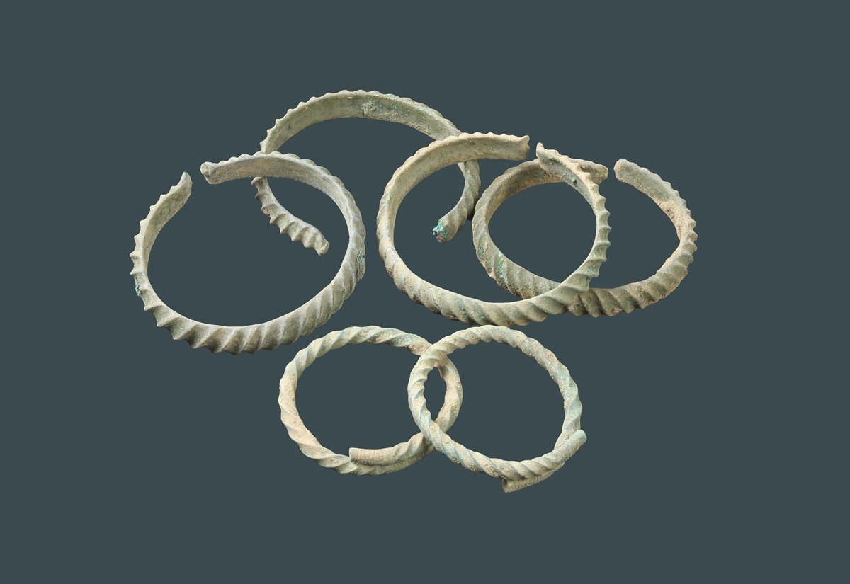Mehrere bronzene Armringe mit gedrehten und glatten Oberflächen liegen auf dunklem, einfarbigem Hintergrund. Die Ringe zeigen unterschiedliche Durchmesser und sind teilweise geöffnet oder leicht deformiert. Die Oberflächen weisen eine grünliche Patina auf, die für Bronze typisch ist.