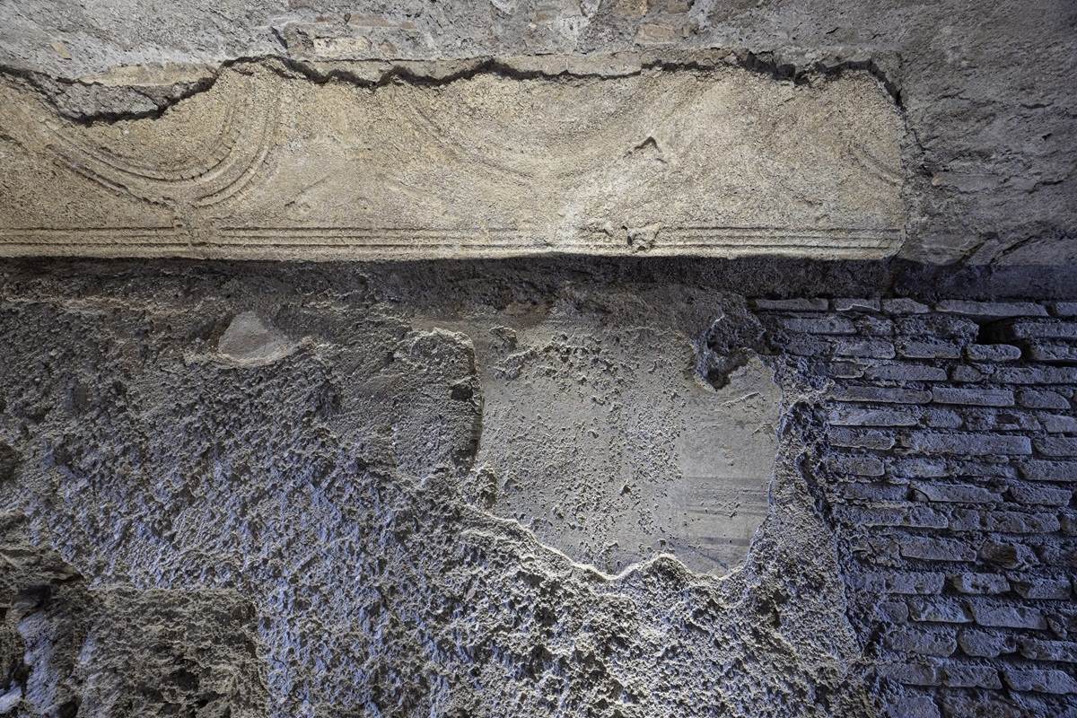 Verwitterte Wand mit grober Putzstruktur und einem horizontalen, verzierten Steinrelief am oberen Rand.