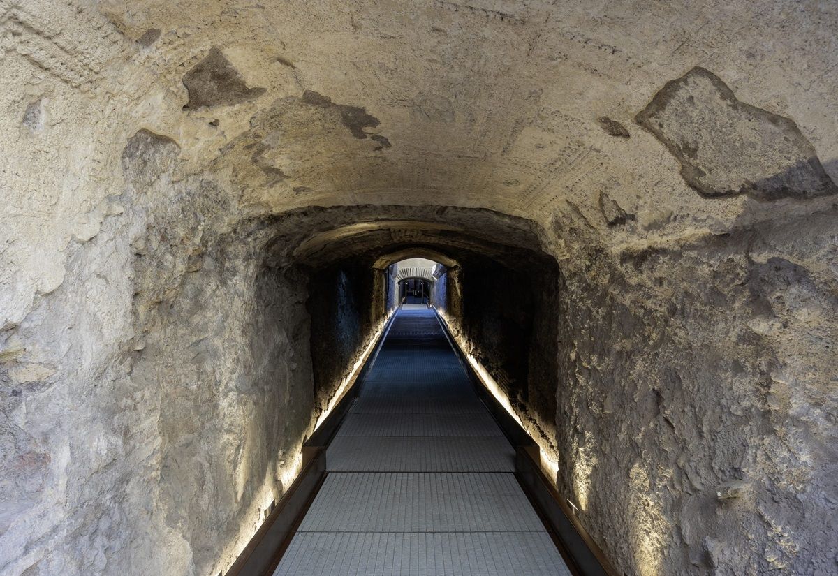 Tunnel mit beleuchtetem Fußweg und rauen Steinwänden, der in der Ferne zu einem helleren Ausgang führt.