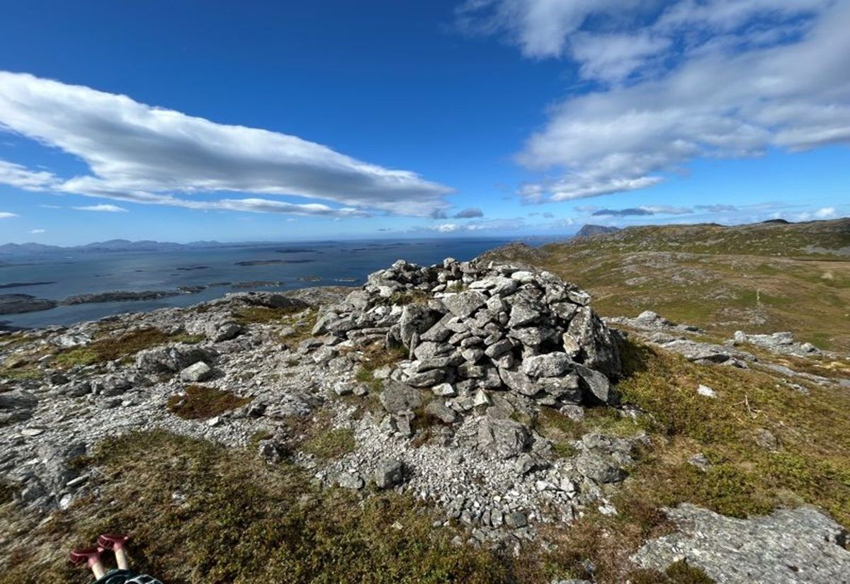 Steinhaufen auf felsigem, moosbewachsenem Hügel unter blauem Himmel mit Wolken und Blick auf Meer und Inseln im Hintergrund.