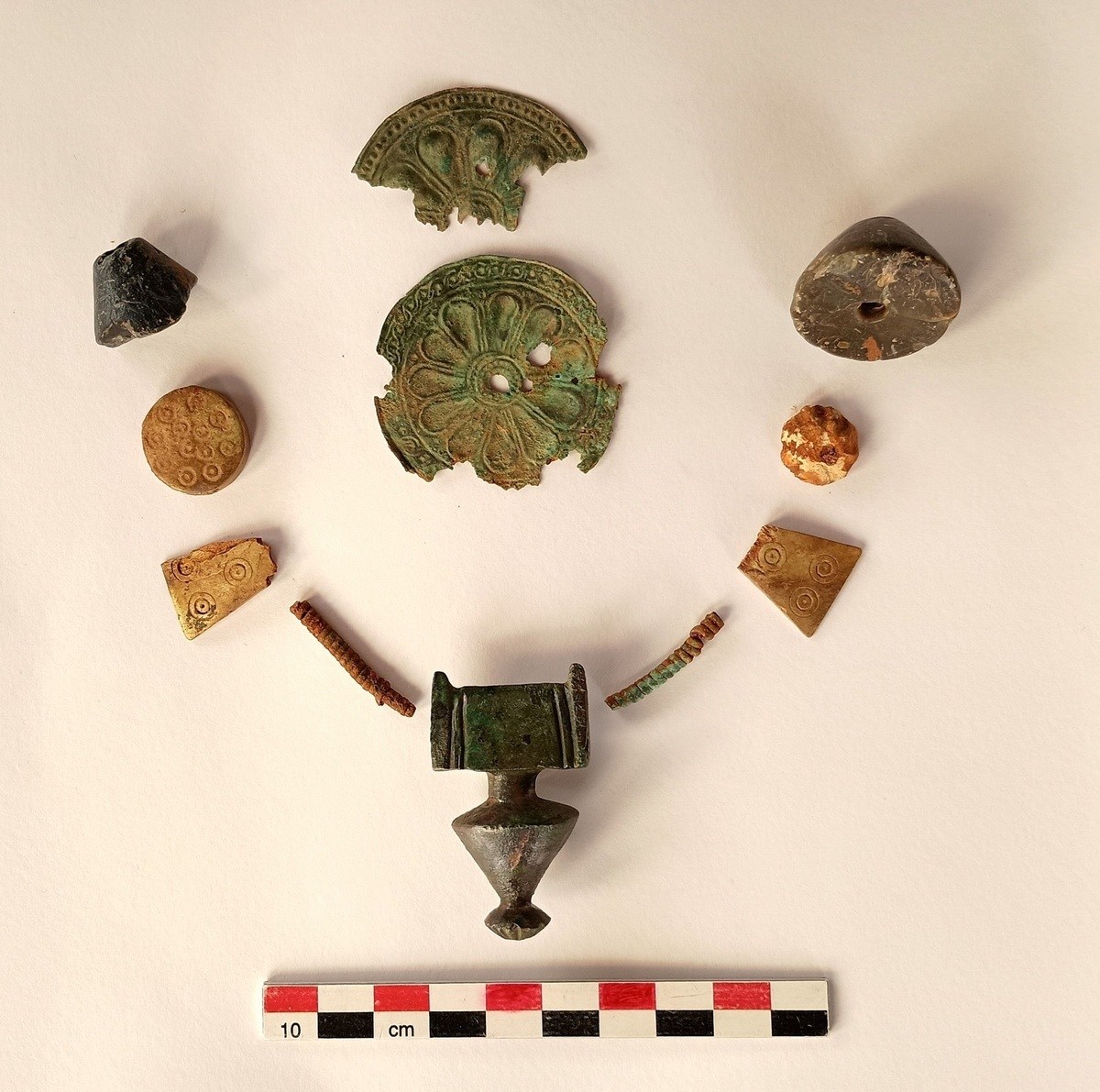 Mehrere archäologische Fundstücke aus Metall und Stein, darunter zwei halbkreisförmige verzierte Metallfragmente, runde und dreieckige Steine mit Mustern, sowie ein zylindrisches Metallobjekt mit kegelförmigem Ende, angeordnet über einem Maßstab mit Zentimeterangaben.