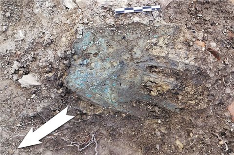 Die Hand von Irulegi bei der Ausgrabung: die Hand aus Bronzeblech liegt korrodiert und dreckig auf der Erde, daneben ein Maßstab und ein Nordpfeil.