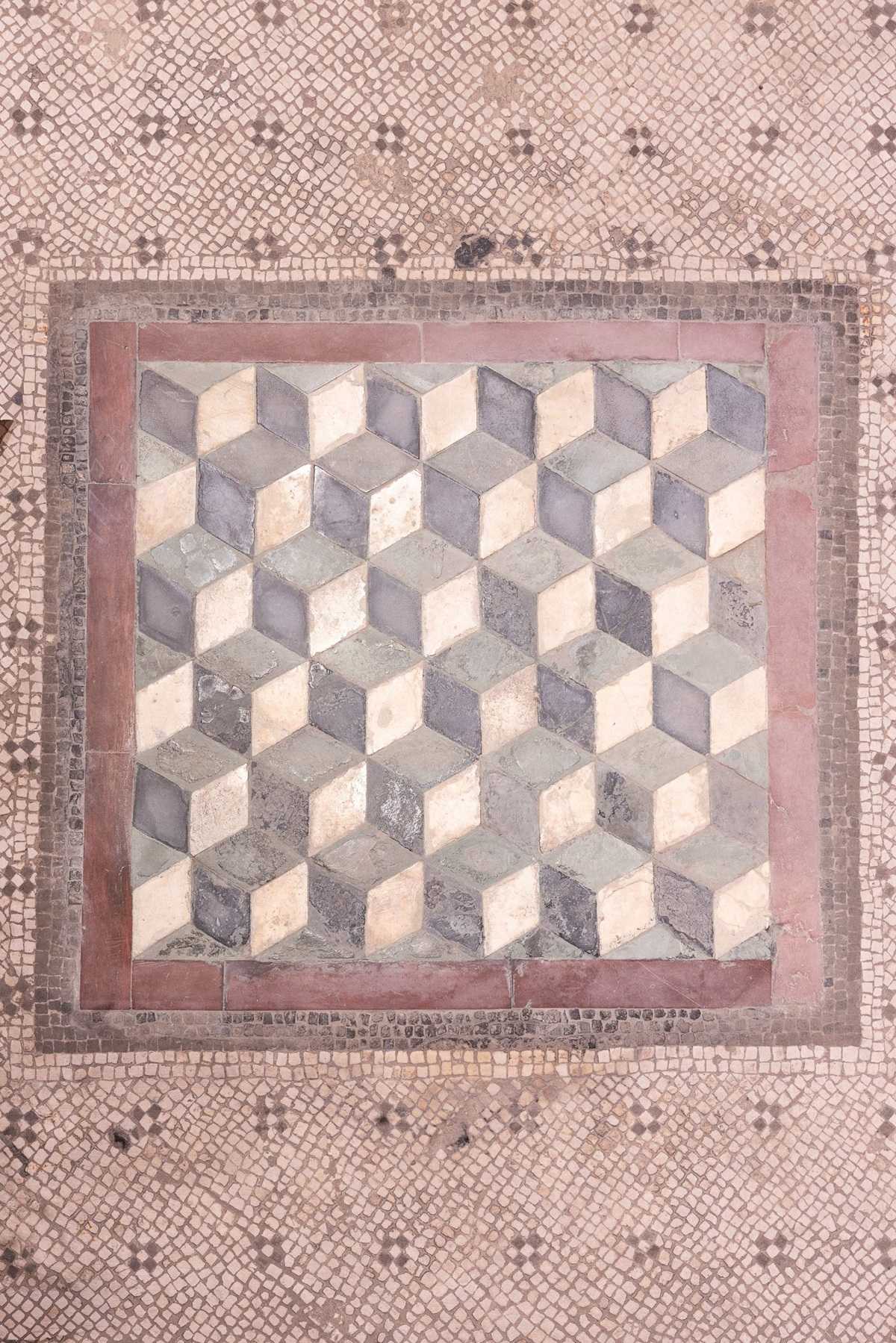 Bodenmosaik mit einem quadratischen Muster aus dreidimensional wirkenden Würfeln, umgeben von einem roten Rahmen und einem Hintergrund aus kleinen, quadratischen Mosaiksteinen.