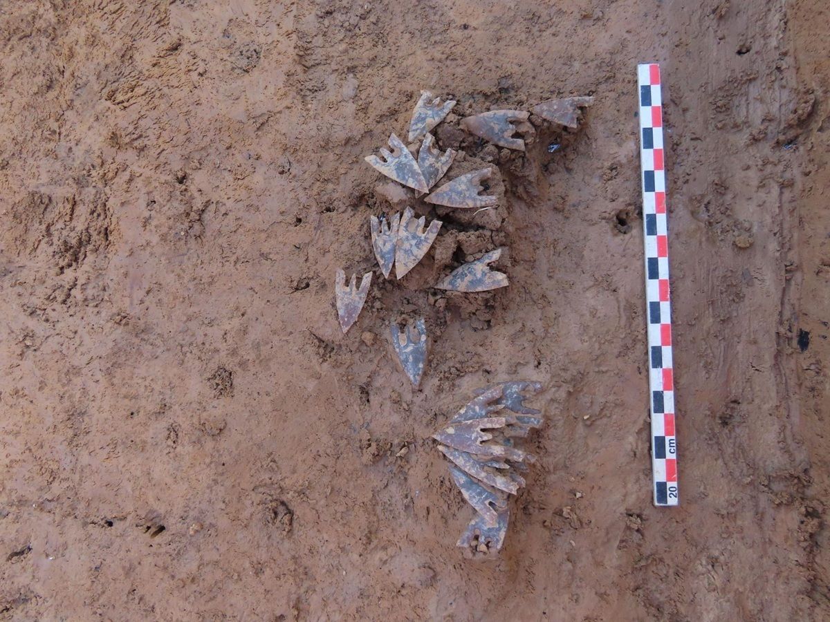 Im Zentrum liegen mehrere dreieckige, flache Objekte aus Metall mit einer Lochung, die auf einem hellbraunen Erduntergrund angeordnet sind. Rechts daneben befindet sich ein Maßstab mit abwechselnd roten und weißen Segmenten.