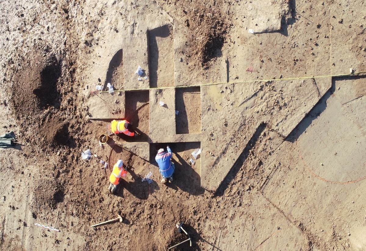 Drei Personen mit Warnwesten arbeiten auf einer Ausgrabungsfläche mit rechteckigen und quadratischen Gruben im hellen Erdreich. Die Fläche ist von einer gelben Messlatte begrenzt. Verschiedene Werkzeuge und kleine Plastiktüten liegen verstreut auf dem Boden.