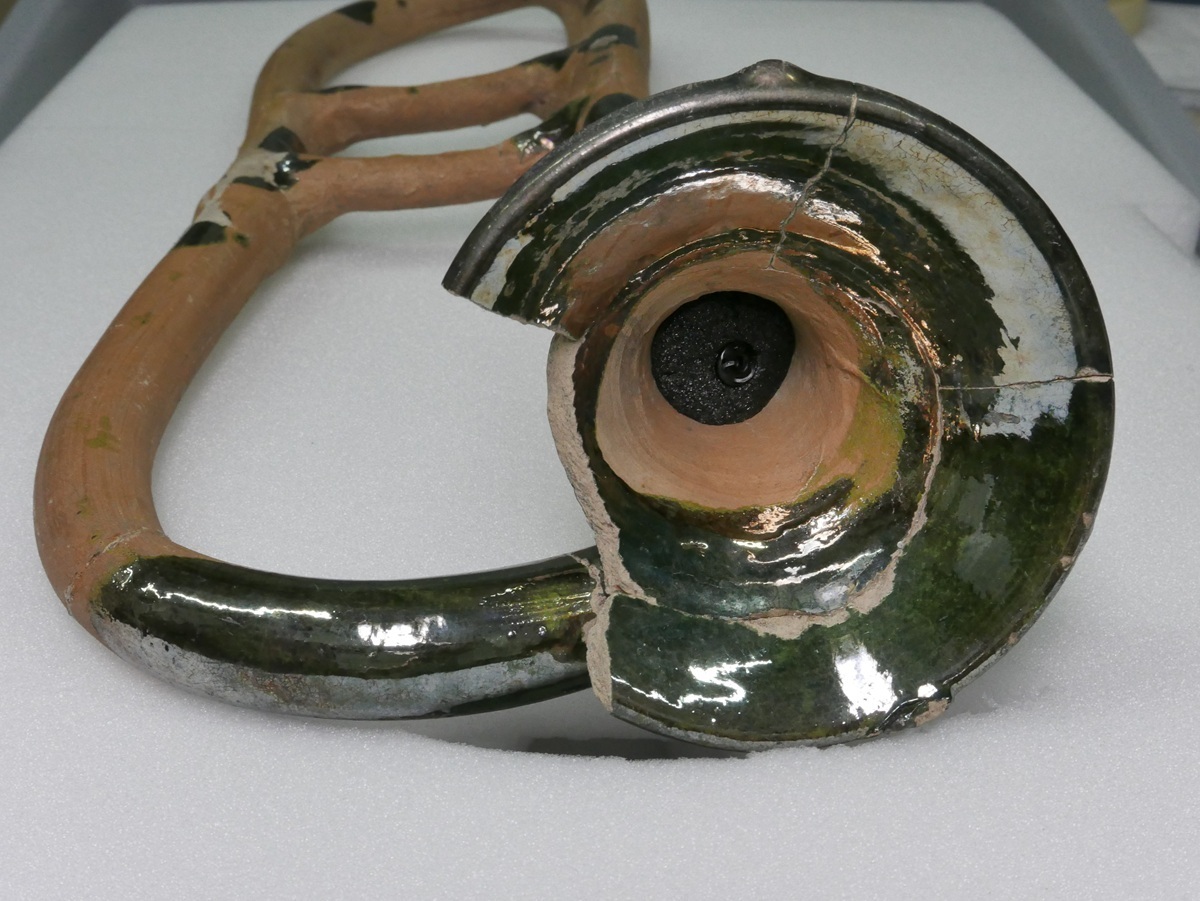 Getöpferte Trompete mit glänzender Glasur, beschädigtem Schalltrichter, liegt auf weißem Untergrund.