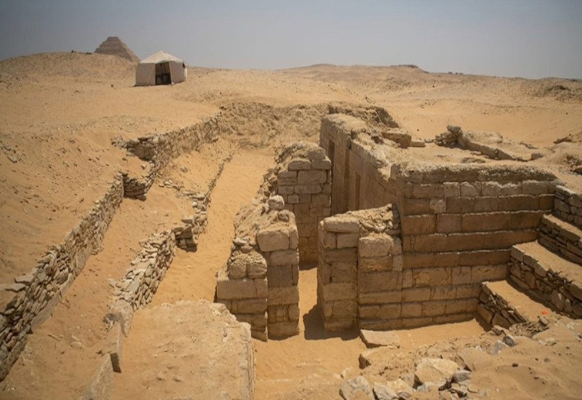 Freigelegte Ruinen aus Sandstein in einer Wüstenlandschaft mit einer kleinen Hütte und einer Pyramide im Hintergrund
