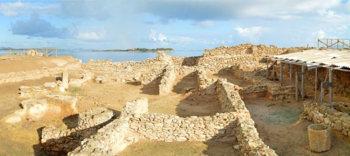 Ruinen einer archäologischen Stätte mit niedrigen Steinmauern und Blick auf das Meer unter bewölktem Himmel