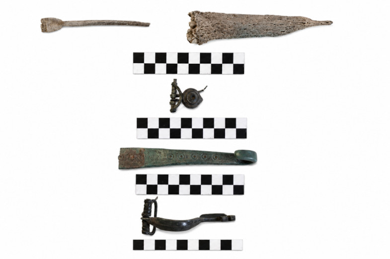 Mehrere archäologische Fundstücke aus der eisenzeitlichen Siedlung am Galgenbichl liegen auf weißem Hintergrund, jeweils neben schwarz-weißen Maßstäben. Oben befindet sich ein länglicher, spitz zulaufender Gegenstand aus Metall, darunter ein kleiner, schmaler Stab. In der Mitte sind eine runde Brosche und ein längliches, grünlich oxidiertes Metallstück zu sehen. Unten liegen zwei weitere Metallobjekte mit gebogenen und filigranen Formen. Die Maßstäbe dienen der Größenbestimmung der Fundstücke.
