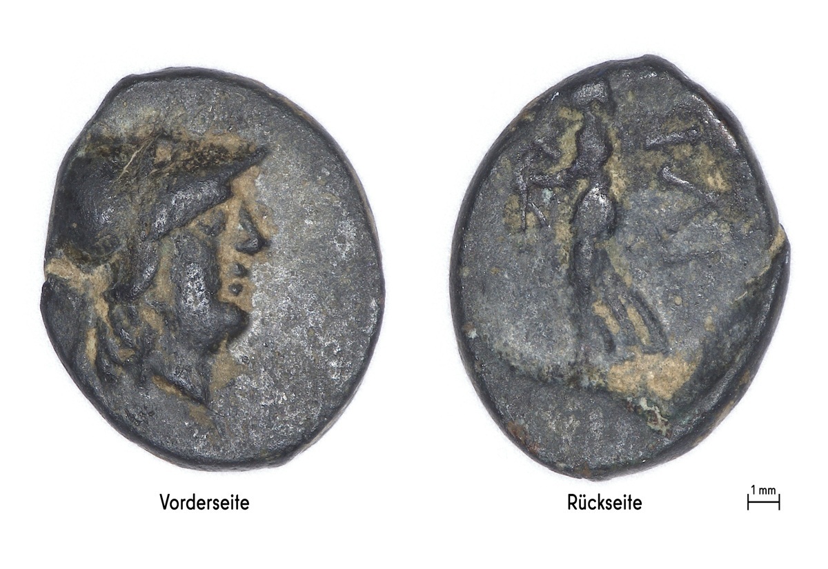 Vorder- und Rückseite einer antiken Münzen werden nebeneinander gezeigt. Links:die Vorderseite zeigt ein Profil eines menschlichen Kopfes mit Helm. Rechts: die Rückseite zeigt eine stehende Figur mit erhobenem Arm. Beide Münzseiten sind dunkel und weisen deutliche Gebrauchsspuren sowie Abnutzungen auf.
