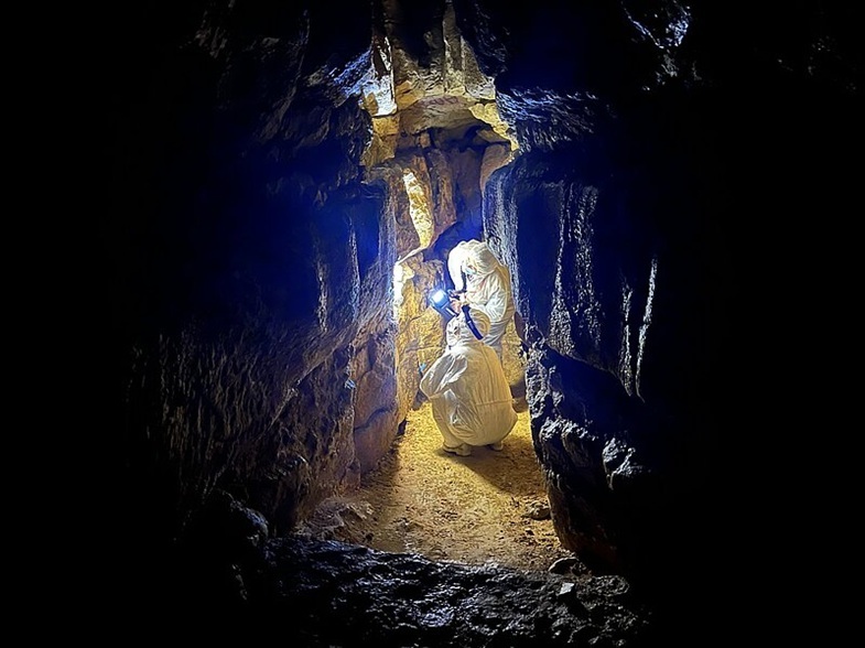 Zwei Personen in einem weißen Schutzanzug. Die eine kniet in einer engen, von Felswänden umgebenen Höhle. Die andere steht in der Höhle und hält eine Taschenlampe und leuchtet damit auf eine Stelle an der Wand. Der Boden ist uneben und mit Staub bedeckt, die Szene wird von einem starken Lichtschein erhellt, der die Struktur der Felsen betont.
