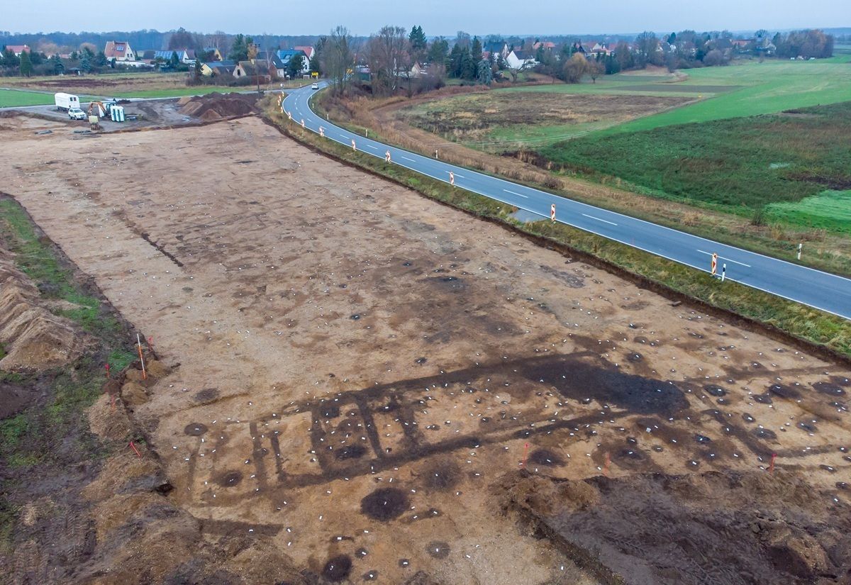 Archäologische Ausgrabungsfläche mit deutlich sichtbaren dunklen Umrissen eines rechteckigen Hausgrundrisses im Erdreich. Zahlreiche kreisförmige Verfärbungen markieren Fundstellen oder Pfostenlöcher. Im Hintergrund verläuft eine Landstraße, daneben Felder und einige Gebäude. Die Aufnahme zeigt die Landschaft aus erhöhter Perspektive.