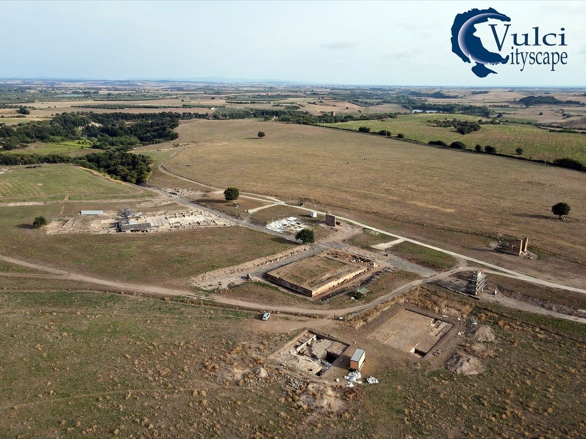 Luftaufnahme einer archäologischen Ausgrabungsstätte in einer weiten, flachen Landschaft mit Feldern und vereinzelten Bäumen. Mehrere rechteckige Ausgrabungsgruben und teilweise freigelegte Mauern sind sichtbar. Im Hintergrund erstreckt sich eine offene, landwirtschaftlich genutzte Fläche bis zum Horizont. Im rechten oberen Bildbereich ist ein Logo mit der Aufschrift 'Vulci cityscape' zu sehen.