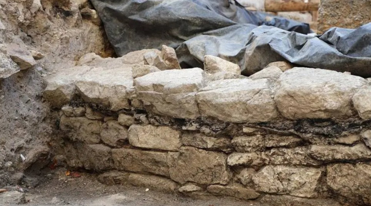 Grobe Steinmauer aus unregelmäßig geformten, unterschiedlich großen Steinen, die ohne Mörtel aufgeschichtet wurden. Die Mauer ist teilweise von Erde umgeben, im Hintergrund liegt eine dunkle Plane über weiteren Ausgrabungsbereichen.