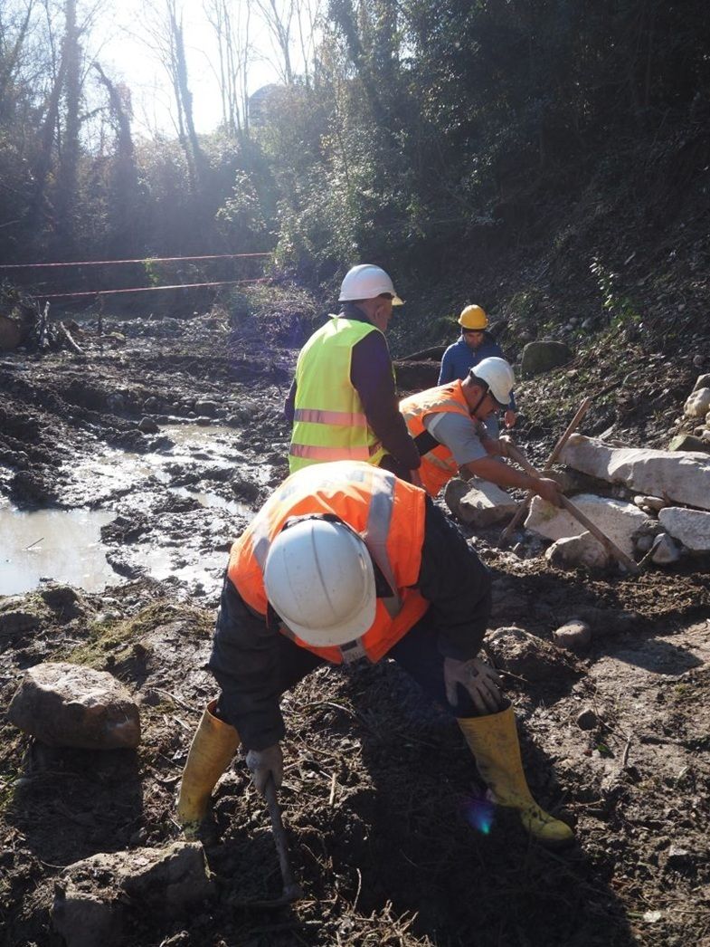 Vier Personen mit Schutzhelmen und Warnwesten arbeiten an einem schlammigen Bachufer. Sie bewegen Steine und Erde mit Werkzeugen. Im Hintergrund sind Bäume und ein kleiner Wasserlauf zu sehen.