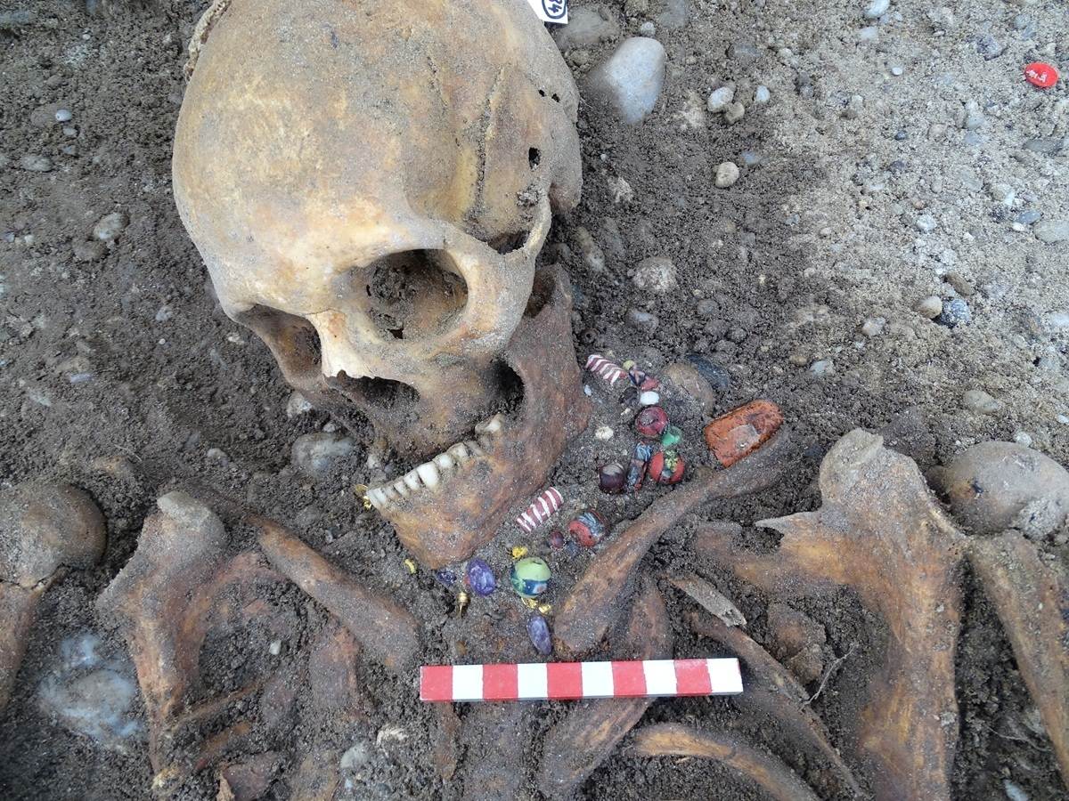 Menschlicher Schädel im Erdreich mit bunten Glasperlen im Halsbereich.