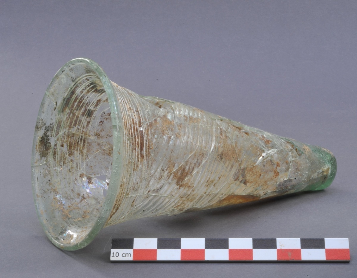 Transparentes, konisches Glasgefäß mit breitem Rand und sichtbaren Gebrauchsspuren, liegt auf der Seite neben einem Maßstab.