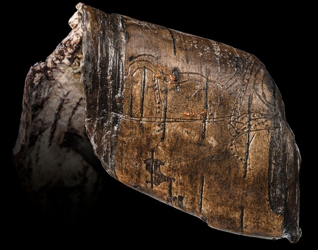 Ein fragmentiertes, braunes Holzobjekt mit eingravierten Linien und Mustern liegt vor schwarzem Hintergrund. Die Oberfläche zeigt deutliche Alters- und Gebrauchsspuren. Die Gravuren verlaufen parallel und kreuzen sich stellenweise, einige Bereiche sind dunkler verfärbt. Das Objekt wirkt unregelmäßig gebrochen und weist an den Kanten helle, raue Stellen auf.