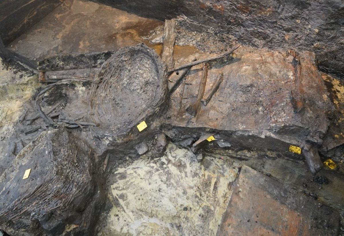 Im Zentrum liegen mehrere dunkle, organische Objekte mit einer geflochtenen Struktur, die an Körbe oder Behälter erinnern. Daneben sind längliche, hölzerne Gegenstände sichtbar, die teilweise von Erde und feuchtem Material umgeben sind. Zwei kleine, gelbe Markierungen sind auf dem Boden platziert. Die Umgebung wirkt feucht und erdig, mit unterschiedlichen Farbtönen von Braun, Grau und Schwarz.