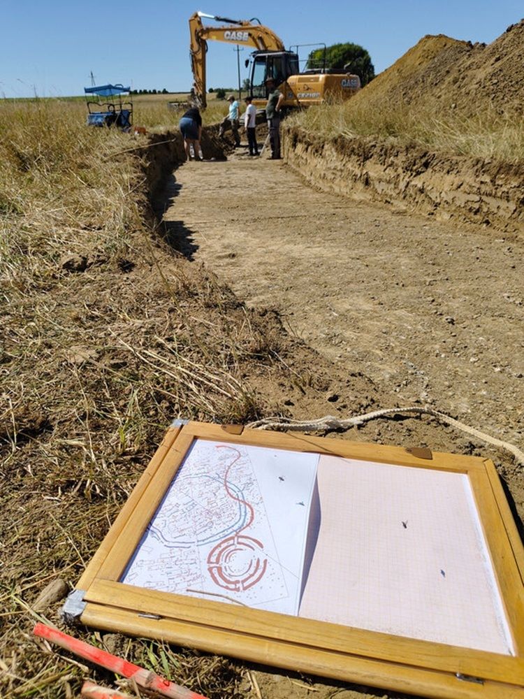 Grabungsschnitt, in dem im hinteren Bereich Meschen stehen. im Vordergrund liegt ein Zeichenbrett, auf dem ein Plan der Grabungsfläche zu sehen ist