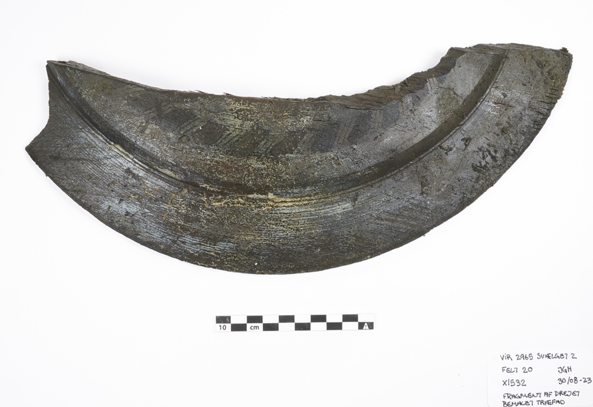 Ein gebogener Gegenstand mit dunkler, teils grünlicher Oberfläche liegt auf weißem Untergrund. Die Oberfläche zeigt eine Reihe von diagonalen Einkerbungen oder Mustern. Unterhalb des Objekts befindet sich eine Maßstabsleiste mit schwarz-weißen Feldern und einer Skala in Zentimetern.