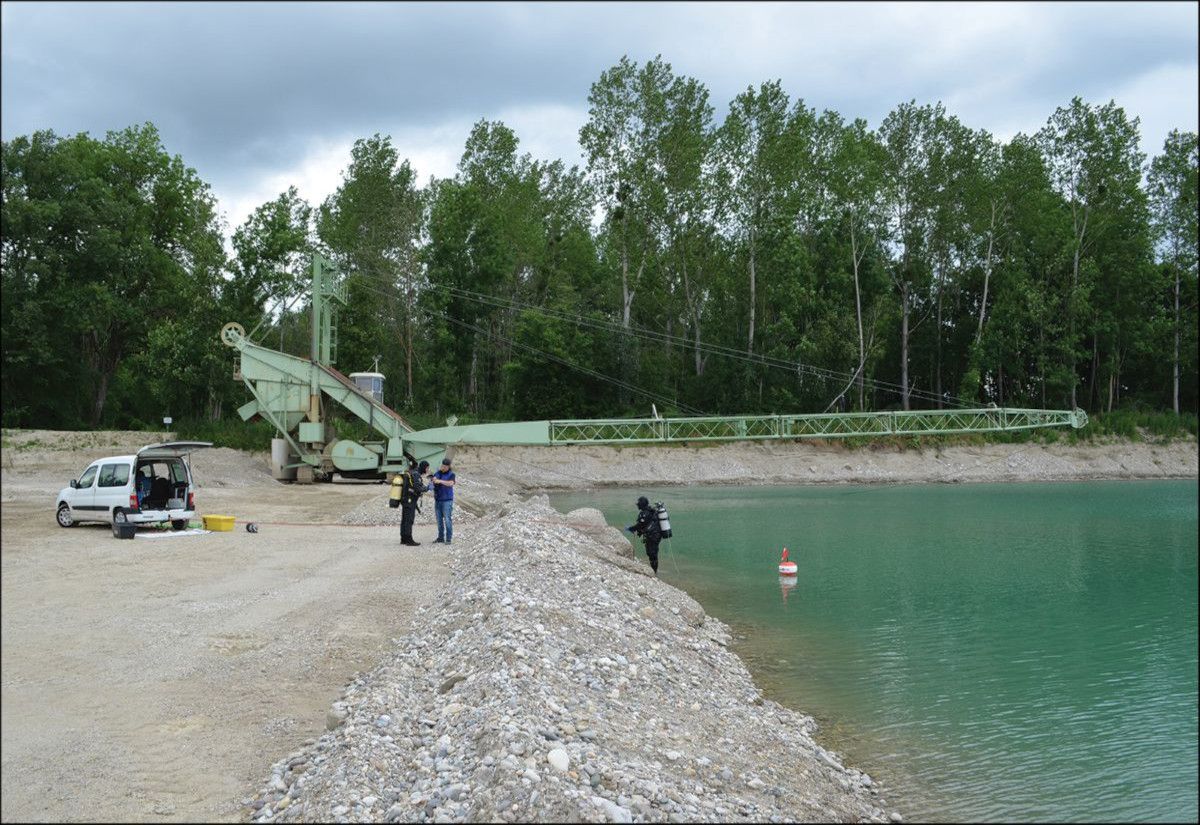 Ein langes, hellgrünes Förderband erstreckt sich schräg über eine Kiesböschung bis zum türkisfarbenen Wasser eines Sees. Am Ufer stehen mehrere Personen neben einem weißen Kleintransporter und technischen Geräten. Europas größter Eisenbarrenfund wurde in Deinham, Oberösterreich gefunden