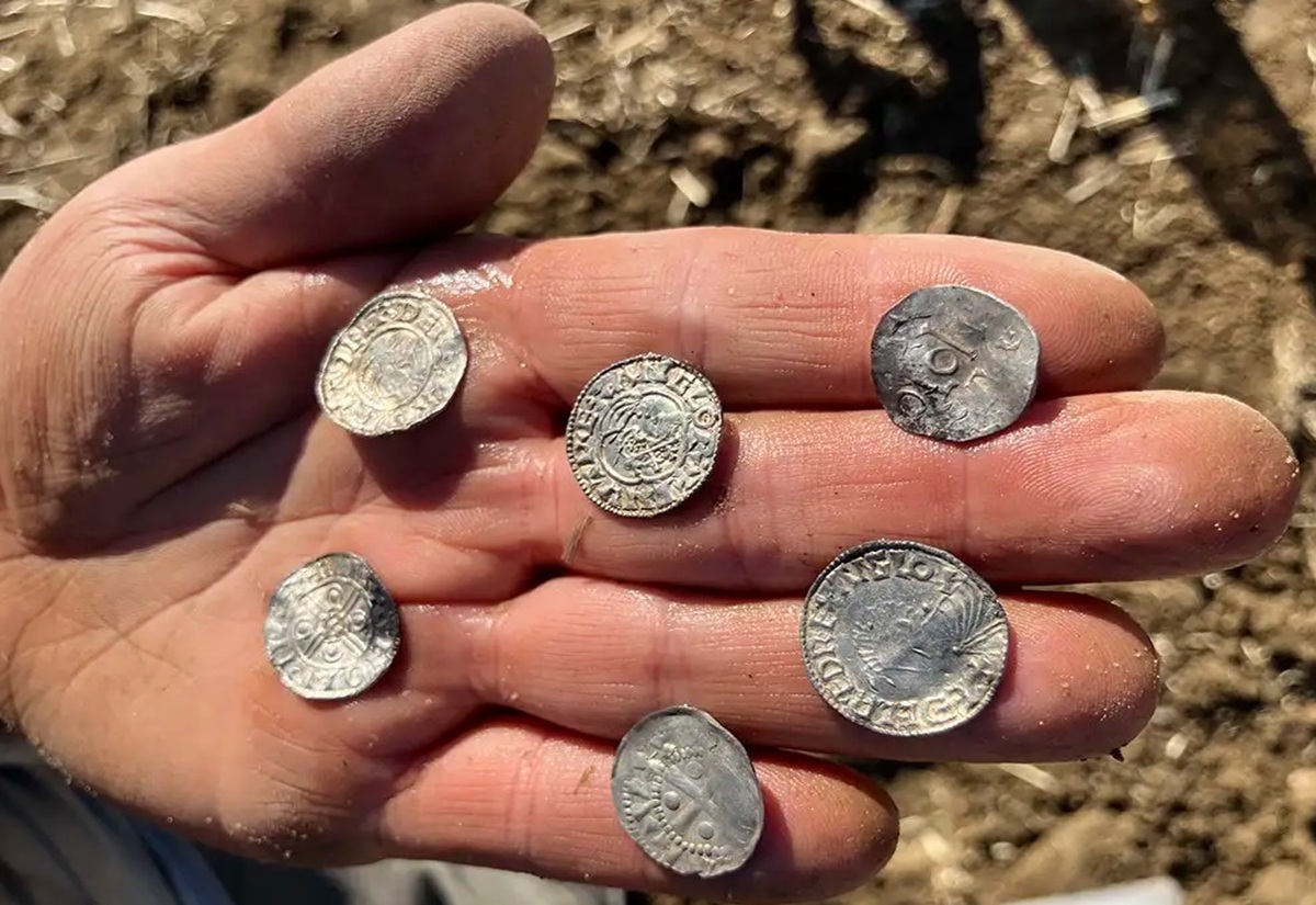 Eine Hand mit erdverschmutzten Fingern hält sechs silberfarbene Münzen. Die Münzen sind unterschiedlich groß und zeigen deutlich sichtbare Prägungen mit kreisförmigen Mustern und Schriftzeichen. Der Hintergrund besteht aus unscharfem, erdigem Boden.