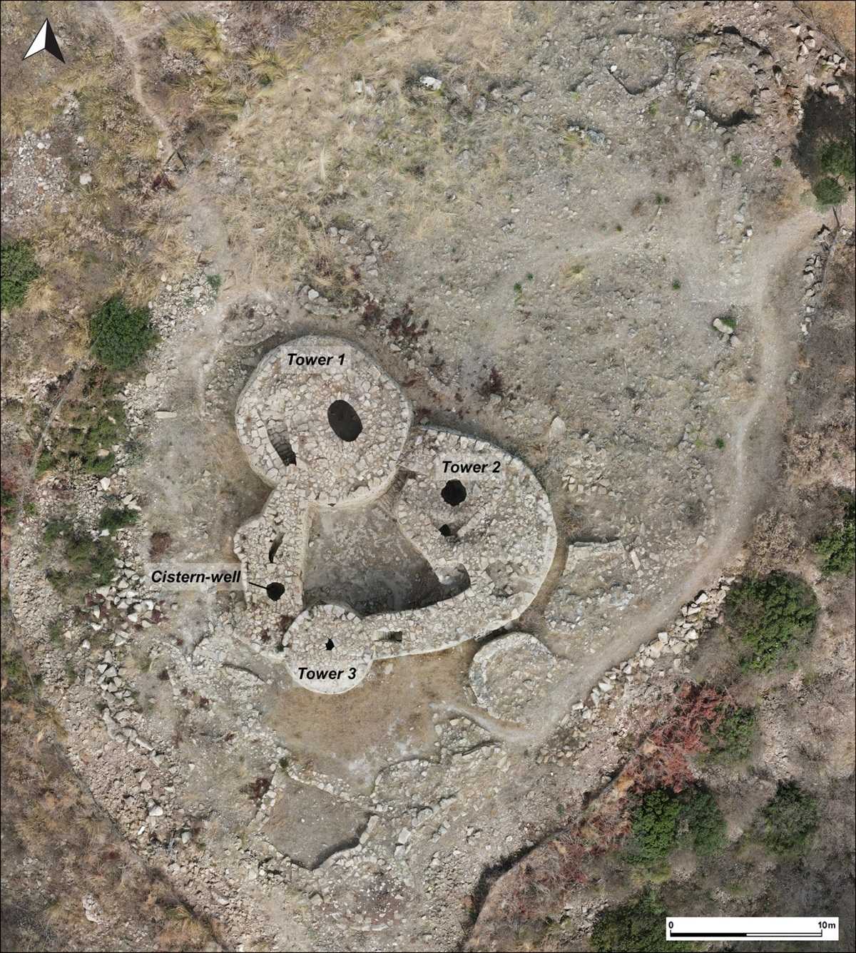 Luftaufnahme einer archäologischen Ausgrabungsstätte mit drei runden Steintürmen, die durch Mauern miteinander verbunden sind. Die Türme sind als 'Tower 1', 'Tower 2' und 'Tower 3' beschriftet. In der Mitte befindet sich eine weitere Markierung mit der Aufschrift 'Cistern-well'. Die Umgebung ist von spärlicher Vegetation und unebenem Gelände geprägt. Ein Maßstabsbalken und ein Richtungspfeil sind am unteren und oberen Bildrand zu sehen.