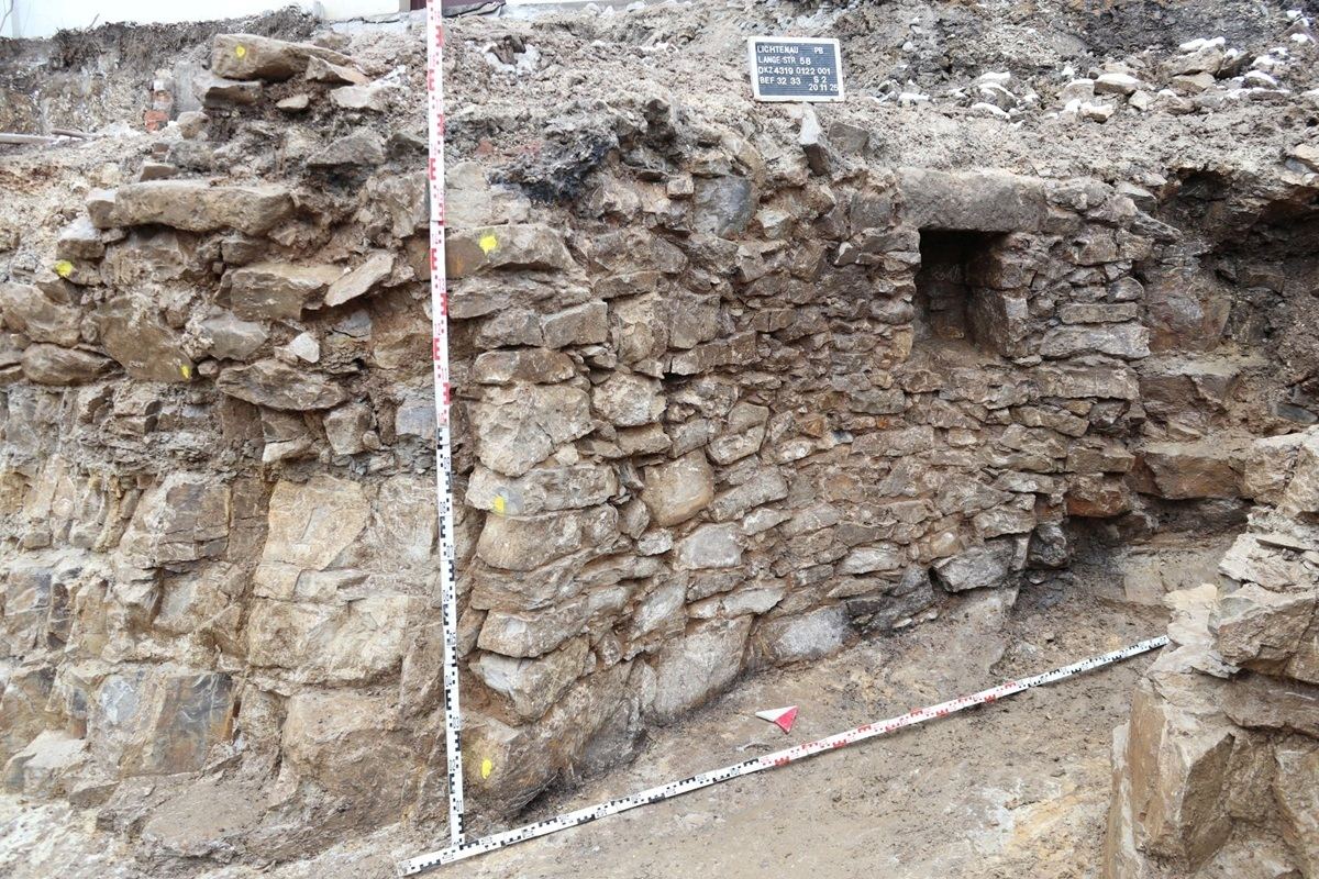 Freigelegter historischer Gewölbekeller. Wand mit unregelmäßig angeordneten, groben Steinen und Mörtel. Die Wand zeigt eine kleine, rechteckige Öffnung. Vor der Wand steht ein Maßstab in L-Form mit Markierungen.