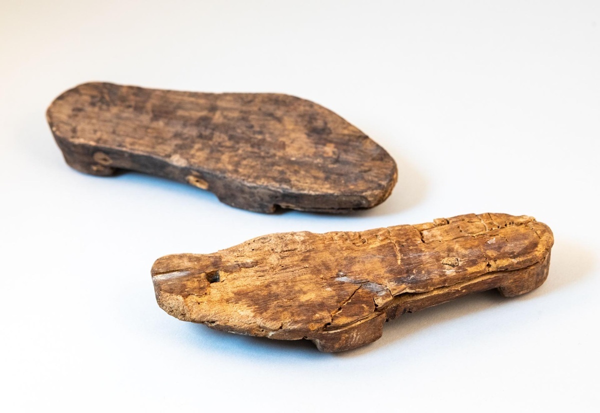 Zwei alte, abgenutzte Holzsohlen mit Nägeln, unregelmäßige Form, auf hellem Untergrund.