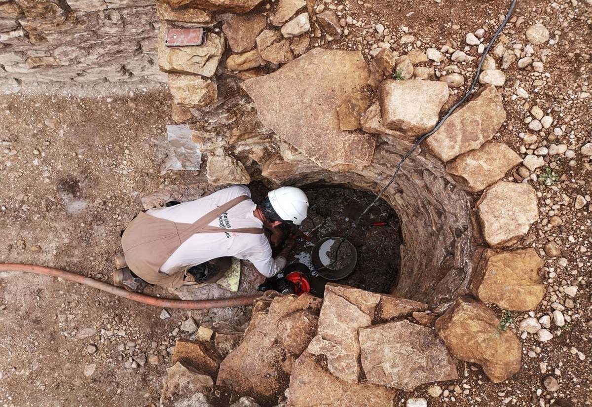 Person mit weißem Helm und weißem Hemd arbeitet in einem runden, steinernen Brunnen, umgeben von Erde und Steinen.